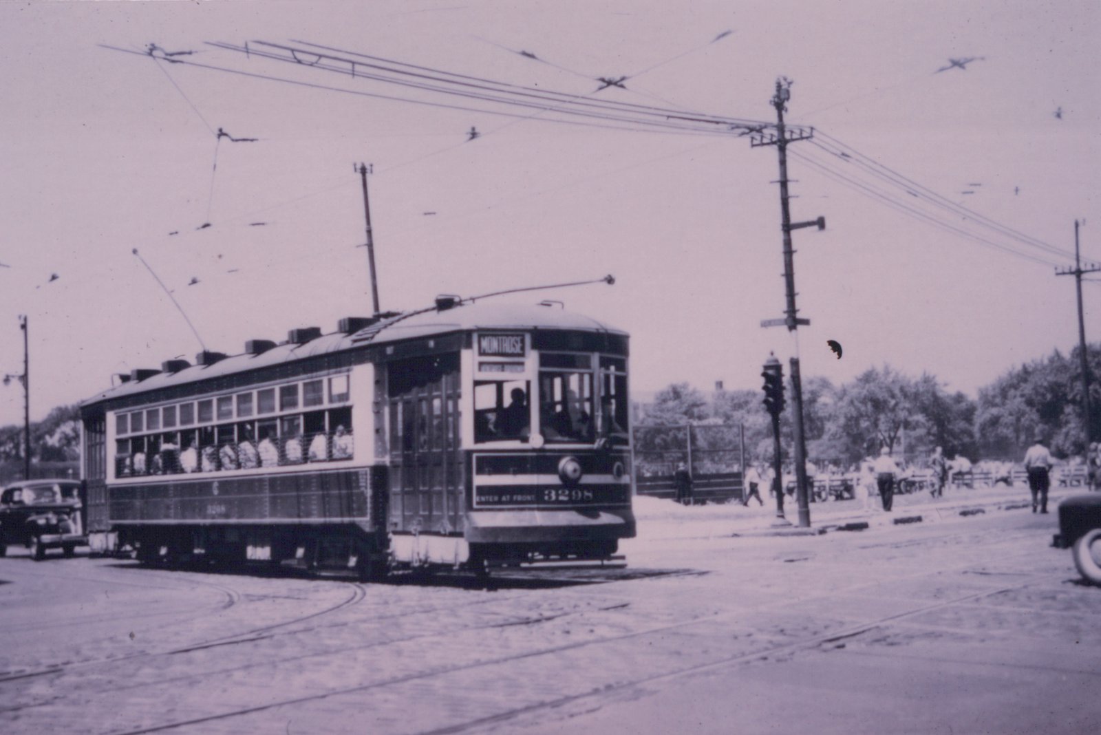 A vintage streetcar with the number 3298 and destination Montrose is shown on a city street with overhead wires, passengers visible inside, and a few pedestrians nearby.