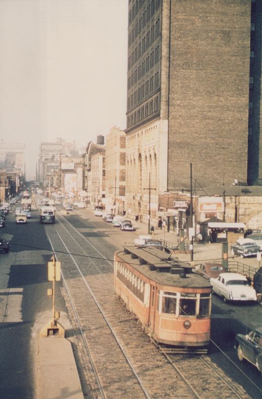 A vintage orange streetcar with the number 75 runs on tracks embedded in a busy, wide city street lined with tall buildings and 1950s automobiles.