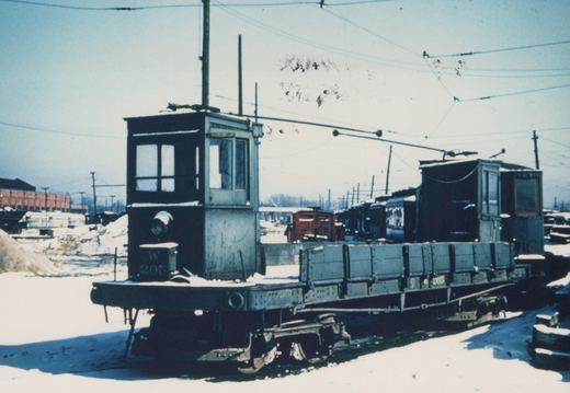 A vintage electric work car with the number W 205 on its front, parked on a snow-covered track in a rail yard with overhead wires and other cars in the background.