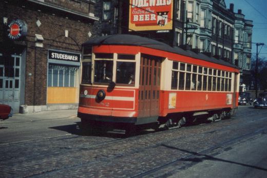 A vintage red streetcar with the number 6152 travels down a cobblestone street lined with brick buildings, including a Studebaker dealership and a Drewrys Beer billboard.