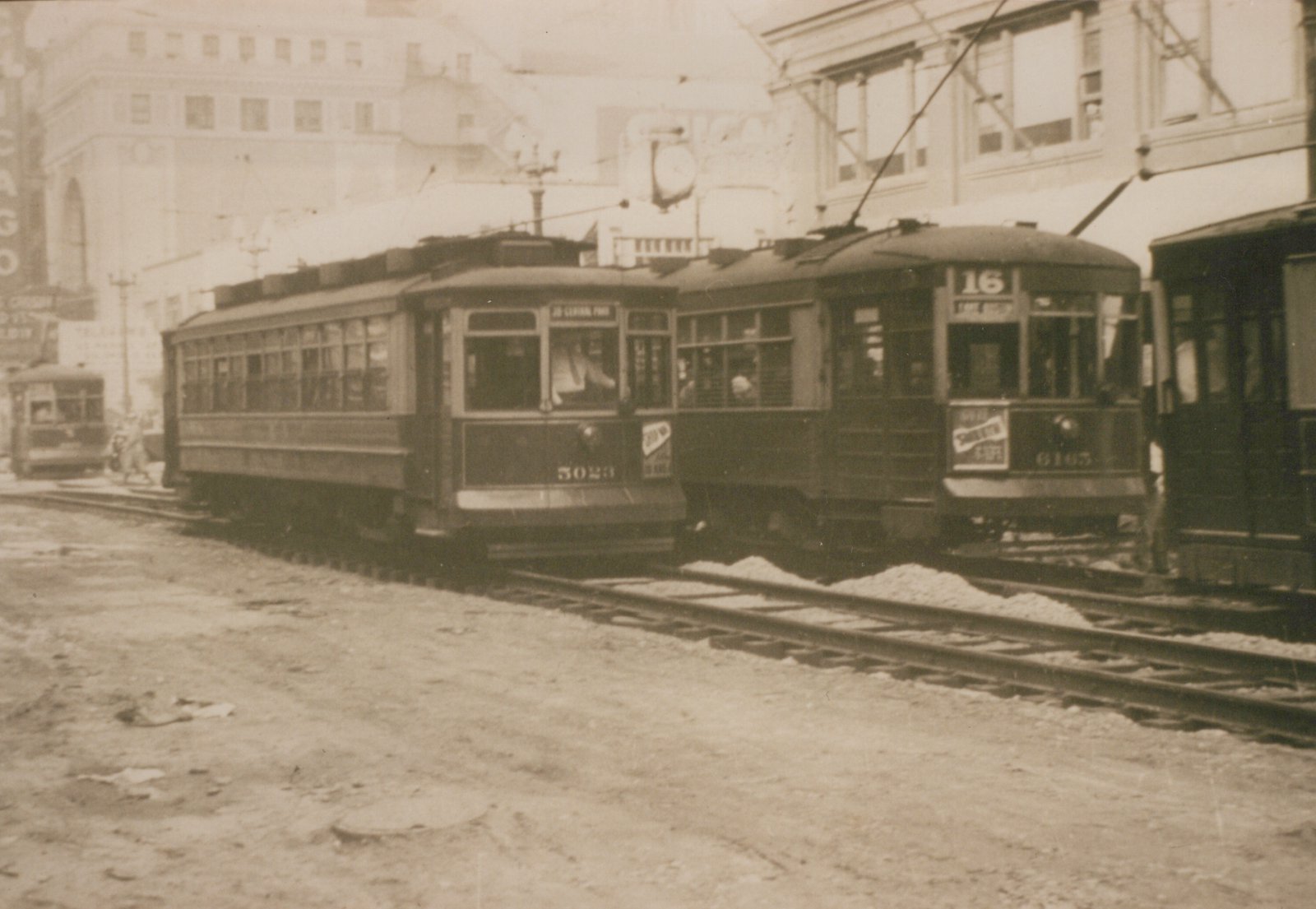 A black and white photograph of two vintage streetcars on a dirt road in Chicago; one is numbered 5023 and the other 6163, both with trolley poles and route signs, set against a backdrop of large buildings.