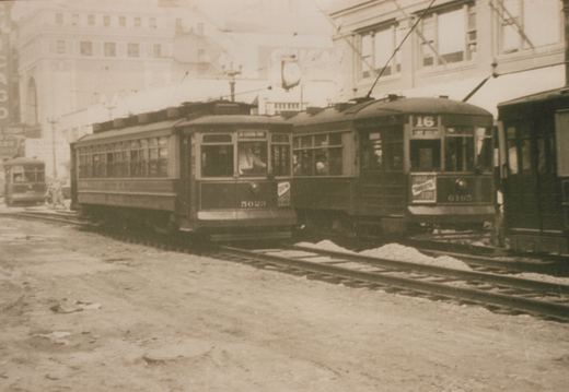 A black and white photograph of two vintage streetcars on a dirt road in Chicago; one is numbered 5023 and the other 6163, both with trolley poles and route signs, set against a backdrop of large buildings.