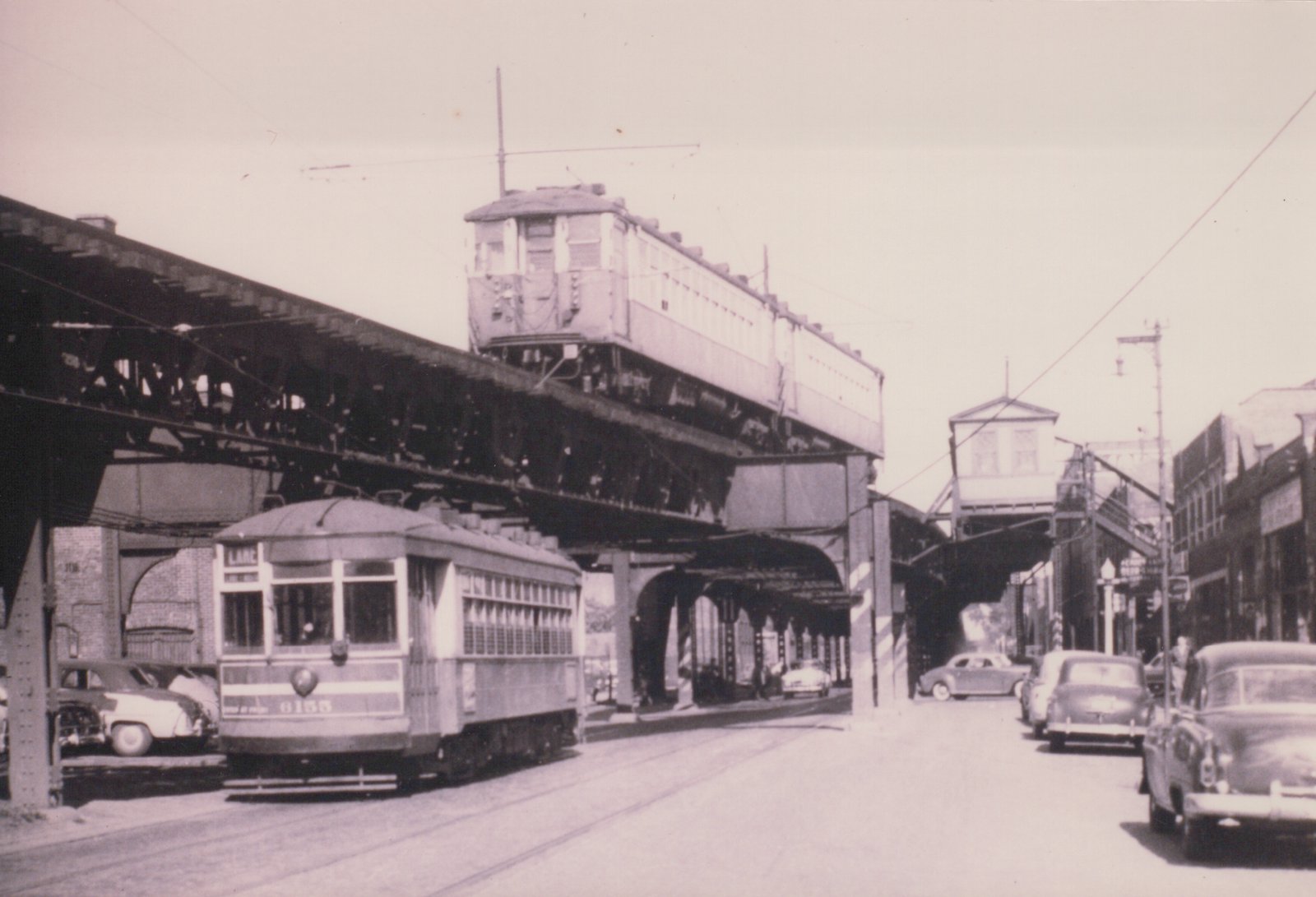 A black and white photo shows a vintage streetcar on a city street with an elevated train structure overhead and several cars parked nearby.