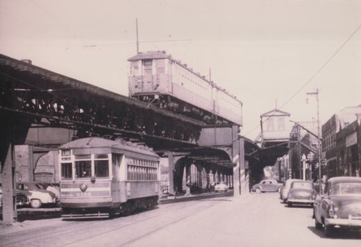A black and white photo shows a vintage streetcar on a city street with an elevated train structure overhead and several cars parked nearby.