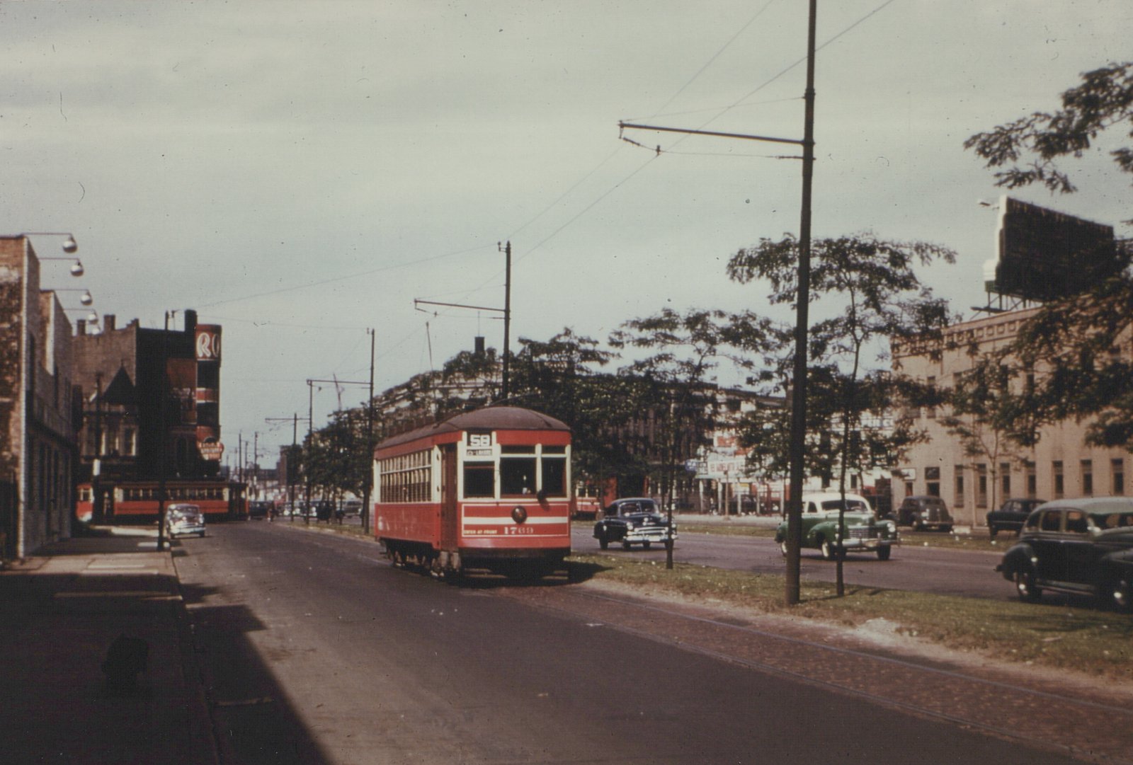 A red and cream vintage streetcar with the number 1769 on the front and route 58 on top, running on tracks down a city street with 1940s cars and buildings.