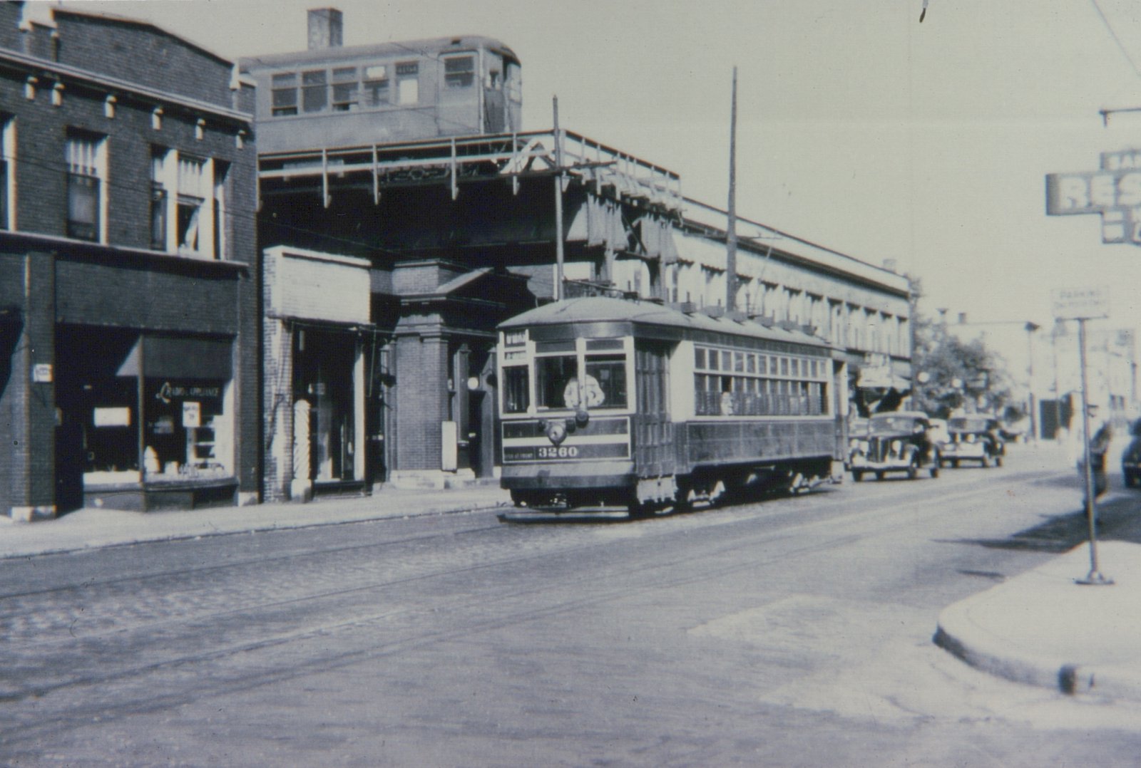 A vintage black and white photo of a streetcar on a city street with an elevated train above it and storefronts along the sidewalk.
