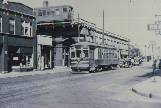 A vintage black and white photo of a streetcar on a city street with an elevated train above it and storefronts along the sidewalk.