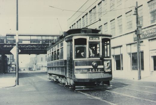 A black and white photograph shows a vintage Chicago streetcar on a city street with an elevated railway overhead and a large building in the background. The car is numbered 1412 and displays the route 26-Cottage.
