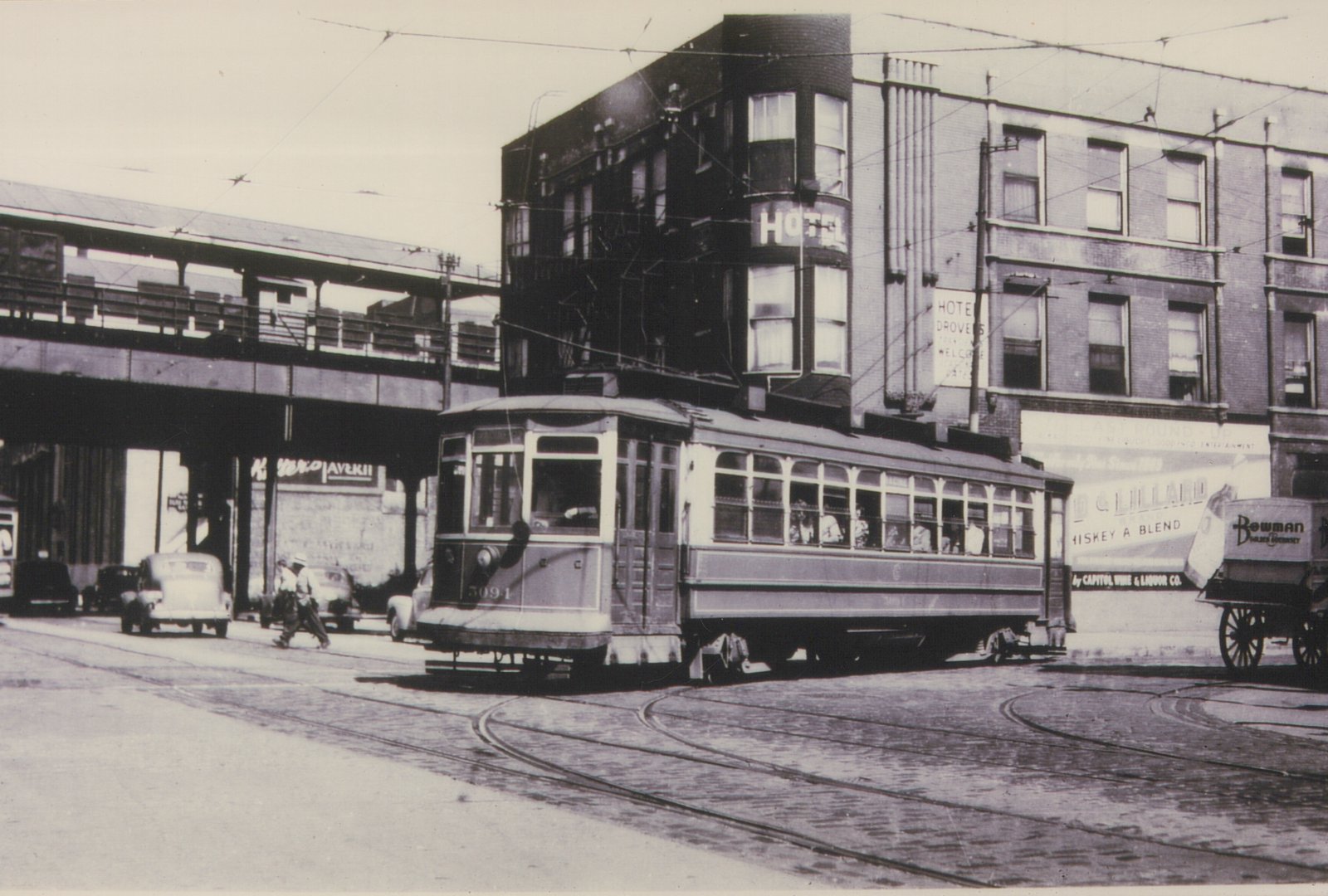 A black and white photograph of a vintage streetcar on a city street, with an elevated train track above and a building with 'HOTEL' signage in the background.