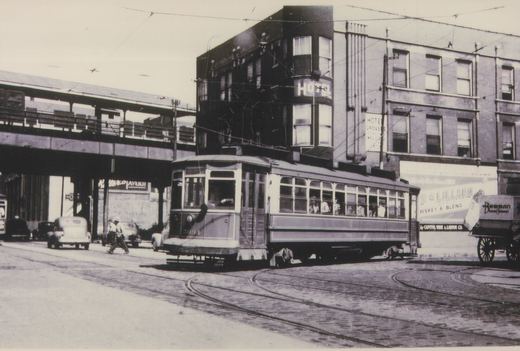 A black and white photograph of a vintage streetcar on a city street, with an elevated train track above and a building with 'HOTEL' signage in the background.