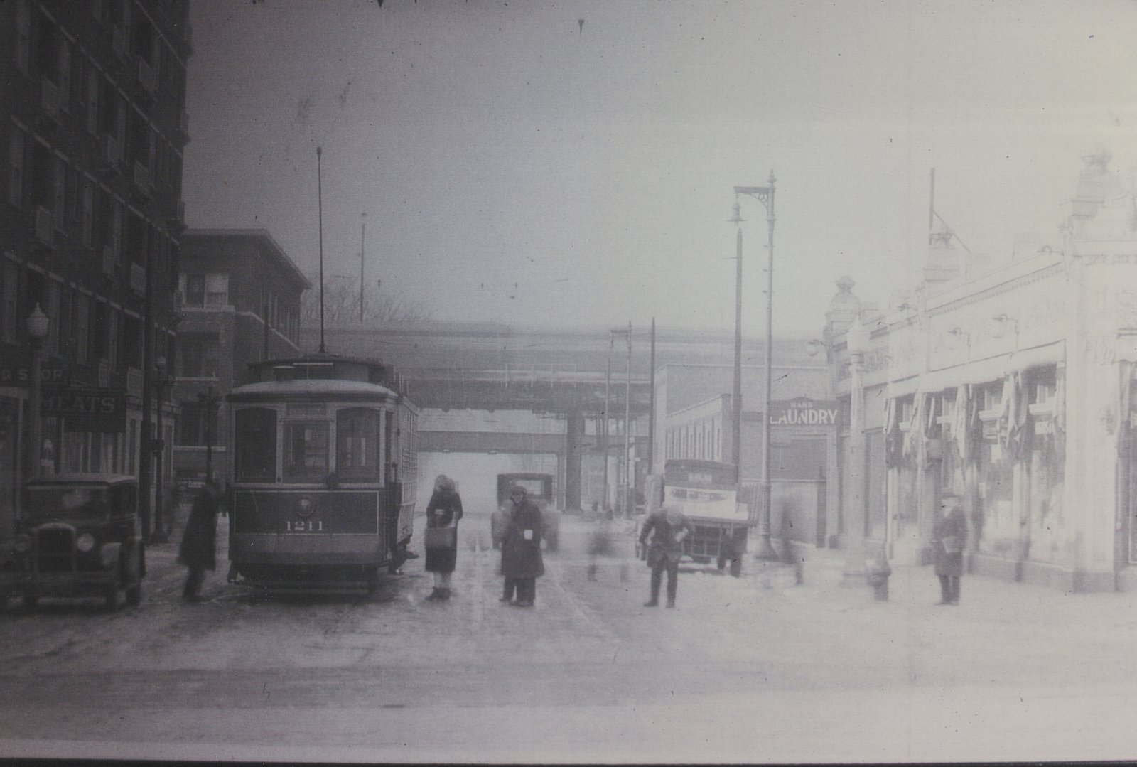 A black and white photograph from the 1930s showing a streetcar on a snowy city street with several people and old cars around it.
