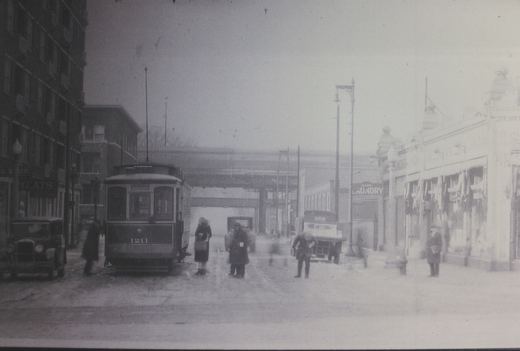 A black and white photograph from the 1930s showing a streetcar on a snowy city street with several people and old cars around it.