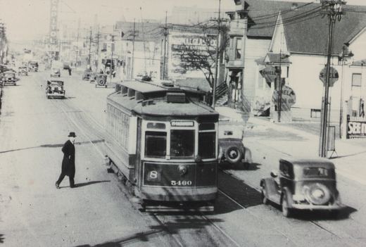 A black and white photo of a vintage streetcar on a city street with a pedestrian crossing and several cars from the era.
