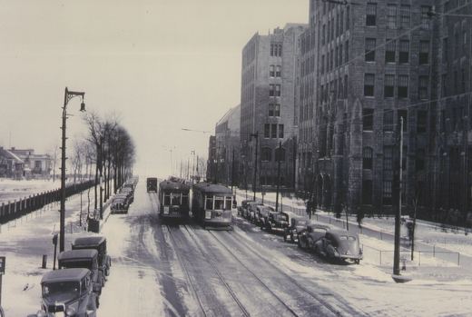 Black and white photo of a snow-covered street with streetcars and parked vintage cars, flanked by large buildings and leafless trees.