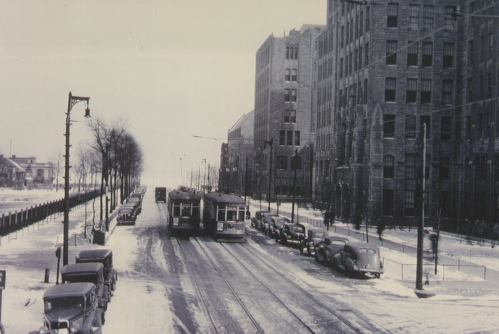 Black and white photo of a snow-covered street with streetcars and parked vintage cars, flanked by large buildings and leafless trees.