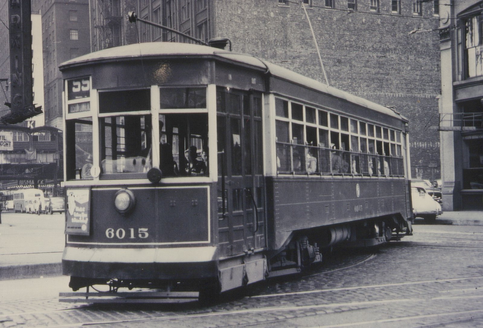 A vintage black-and-white photo of a streetcar with the number 6015 on its front and side, showing passengers inside, on a cobblestone street with buildings and other vehicles in the background.