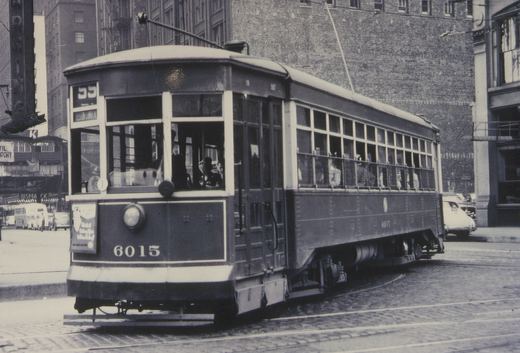 A vintage black-and-white photo of a streetcar with the number 6015 on its front and side, showing passengers inside, on a cobblestone street with buildings and other vehicles in the background.