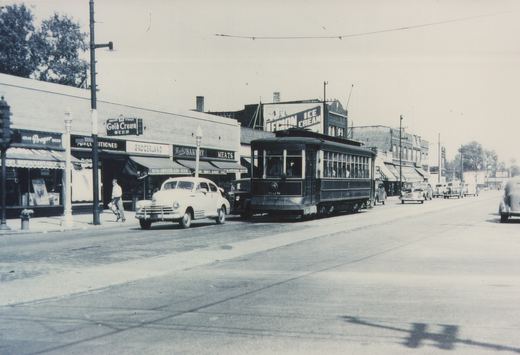 A black-and-white photo of a 1940s streetcar on a city street with vintage automobiles and storefronts.