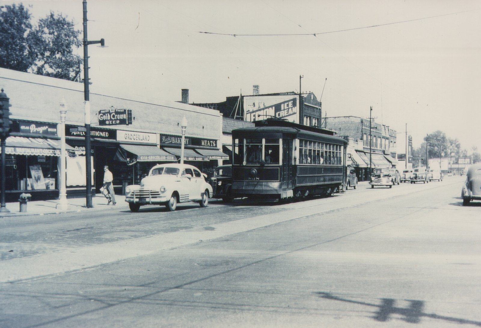 A black-and-white photo of a 1940s streetcar on a city street with vintage automobiles and storefronts.