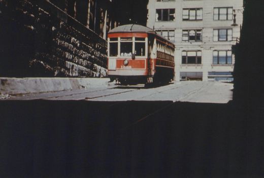 A red and cream streetcar with the number 1608 on its front is stopped on a paved street next to a stone wall and a multi-story building.