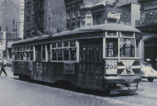 A vintage black-and-white photo of a dark, riveted steel streetcar with the number 3359 and signs for route 4 and Lincoln Park Zoo, stopped on a cobblestone street with a Coca-Cola sign in the background.