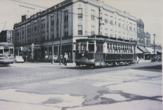 Black-and-white photo of two vintage streetcars at a city intersection, one labeled 604 and the other 8010, with a storefront reading 'Marianne's DRESSES' in the background.