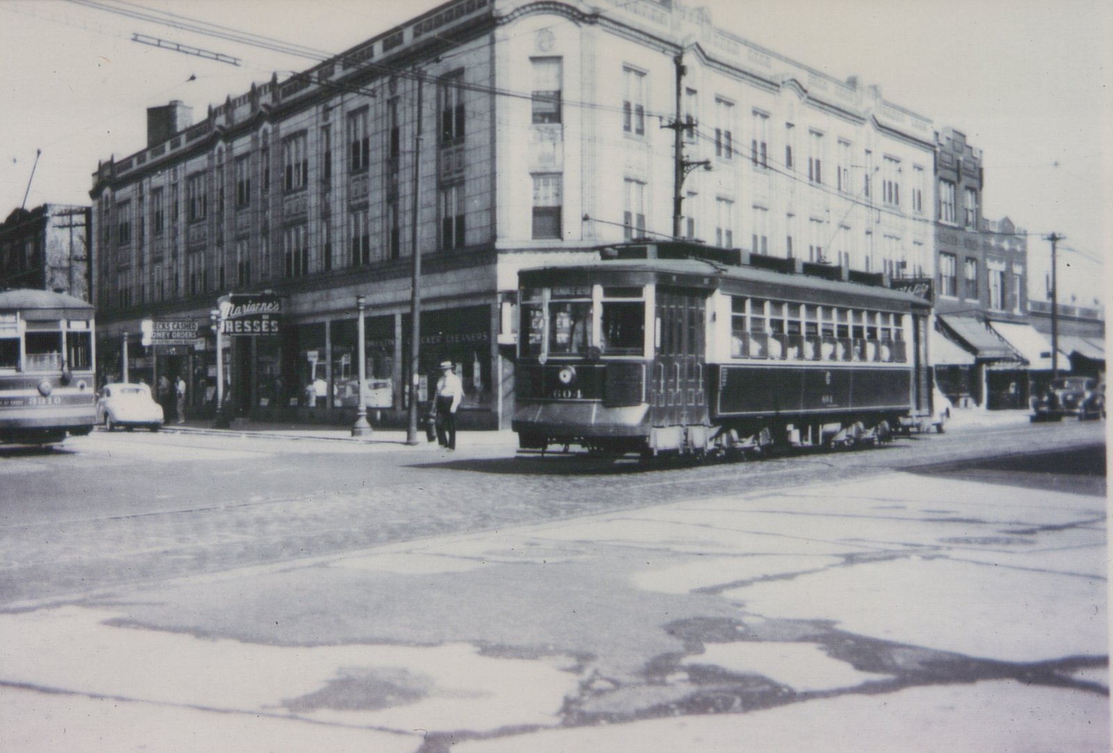 Black-and-white photo of two vintage streetcars at a city intersection, one labeled 604 and the other 8010, with a storefront reading 'Marianne's DRESSES' in the background.
