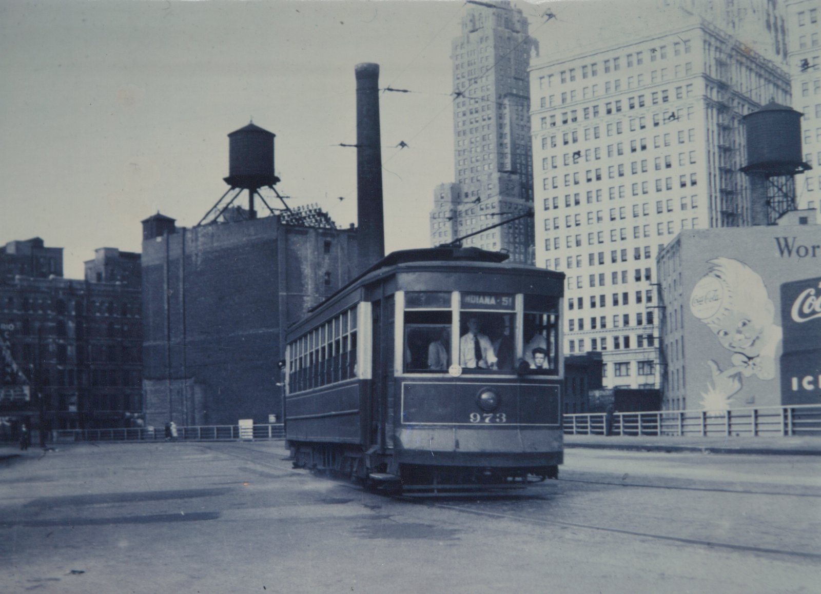 A black-and-white photo of a vintage streetcar with the number 973 on its front, traveling on a city street with tall buildings and a large Coca-Cola billboard in the background.