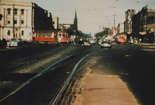 Two red streetcars and vintage cars on a cobblestone street in Chicago, Illinois, in June 1951, with overhead wires and buildings in the background.