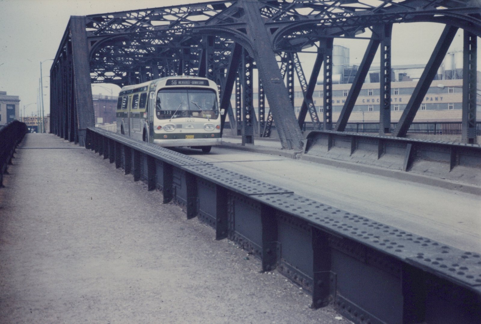 A white and green CTA bus drives across a steel truss bridge on a cloudy day, with a chocolate factory visible in the background.