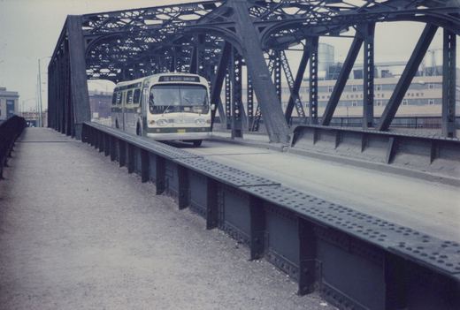 A white and green CTA bus drives across a steel truss bridge on a cloudy day, with a chocolate factory visible in the background.