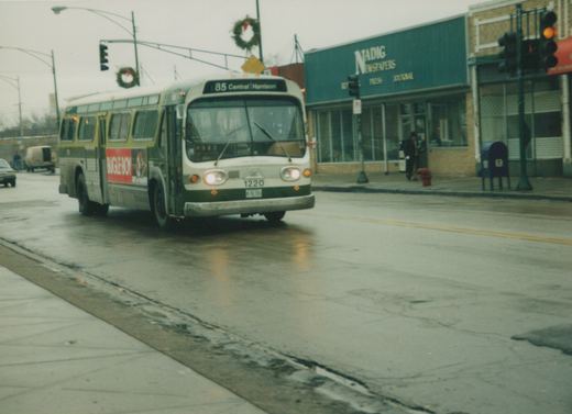 A green and cream Chicago Transit Authority bus, number 1220, is driving on a wet street with holiday wreaths on the lampposts and a newspaper store on the corner.