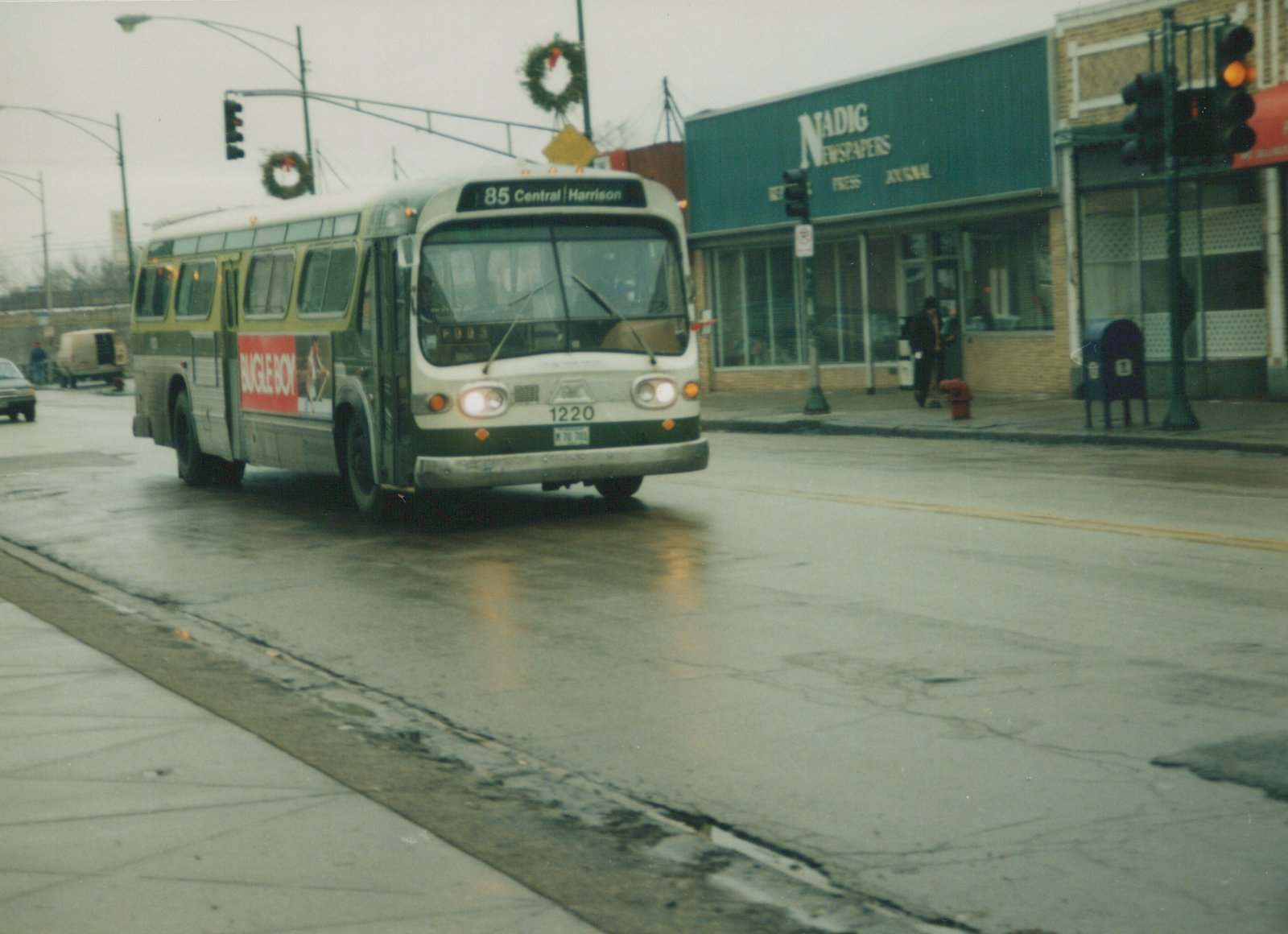A green and cream Chicago Transit Authority bus, number 1220, is driving on a wet street with holiday wreaths on the lampposts and a newspaper store on the corner.