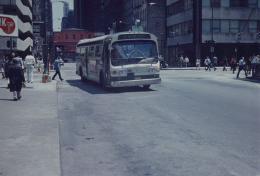 A white and green CTA bus with the number 1357 on the front is turning at a city intersection with pedestrians crossing the street and tall buildings in the background.