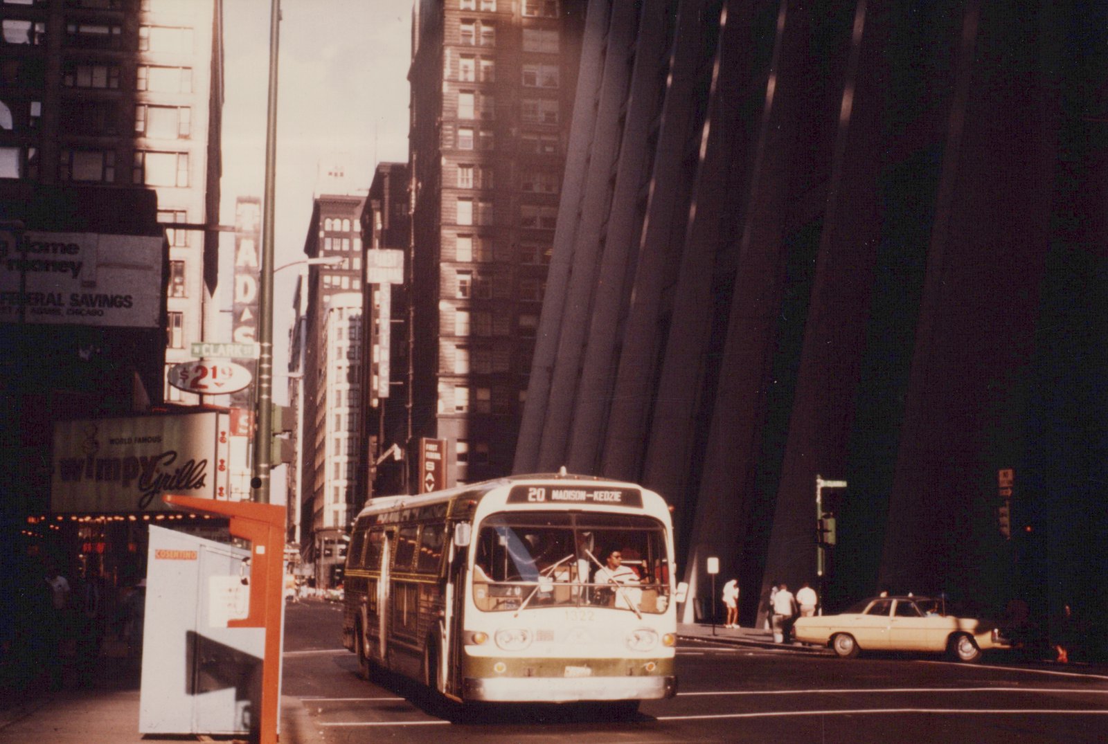 A white and green Chicago Transit Authority bus is parked on a city street, with tall buildings, including the John Hancock Center, and a yellow taxi in the background.
