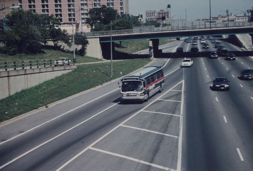 A Chicago Transit Authority bus with a 'Promote Chicago' sign drives on a multi-lane highway under an overpass on a sunny day.