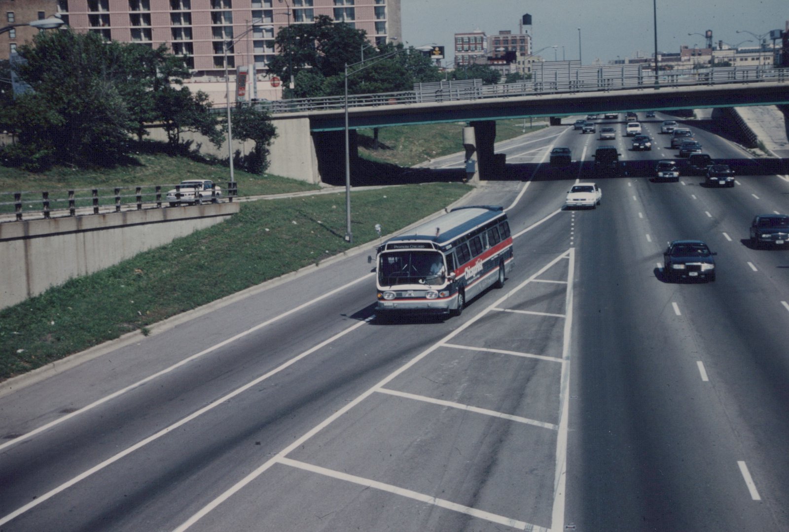 A Chicago Transit Authority bus with a 'Promote Chicago' sign drives on a multi-lane highway under an overpass on a sunny day.