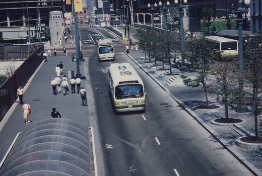 An elevated view of a white and green Chicago Transit Authority bus, number 1262, driving down a city street with pedestrians on the sidewalk and another CTA bus parked nearby.