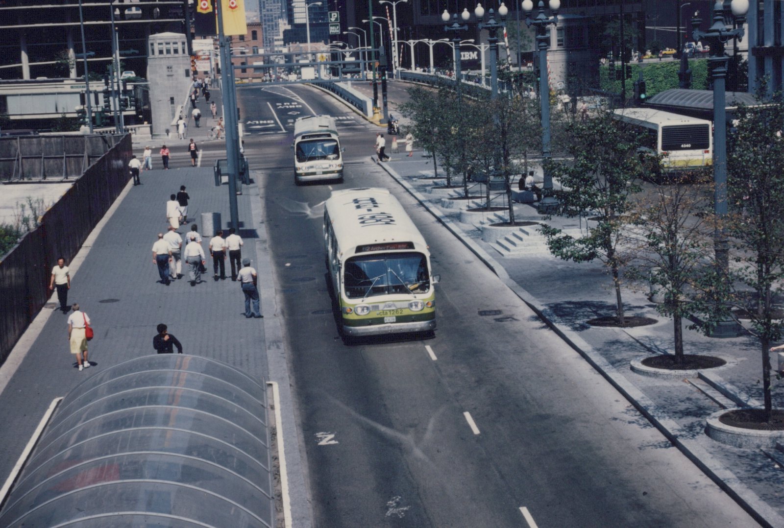 An elevated view of a white and green Chicago Transit Authority bus, number 1262, driving down a city street with pedestrians on the sidewalk and another CTA bus parked nearby.