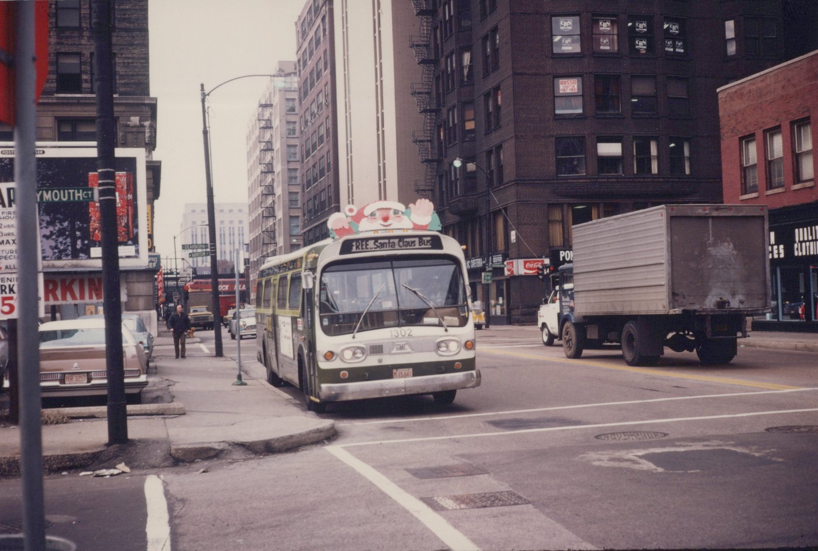 A white and green motor bus with a Santa Claus decoration on its roof, labeled 'FREE...Santa Claus Bus', is stopped on a city street with a pedestrian on the sidewalk and tall buildings in the background.