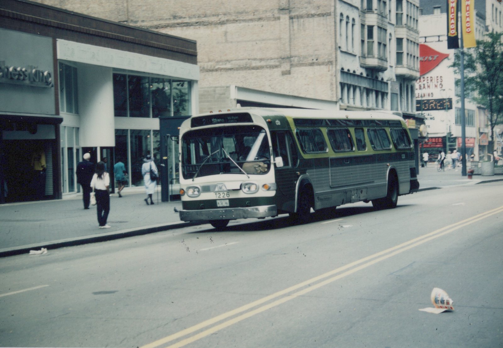 A green, black and white CTA bus numbered 1226 is stopped on a city street with pedestrians on the sidewalk and storefronts including 'Chess King' in the background.
