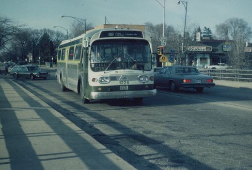 A green and cream CTA bus numbered 1226 is driving on a city street on a clear winter day, with a sedan beside it and bare trees in the background.