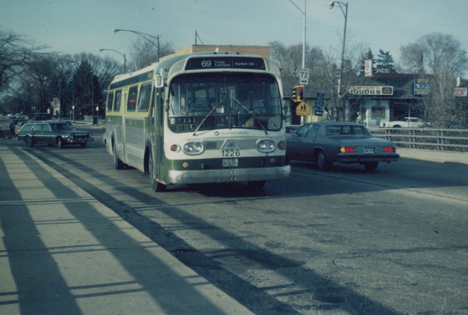 A green and cream CTA bus numbered 1226 is driving on a city street on a clear winter day, with a sedan beside it and bare trees in the background.