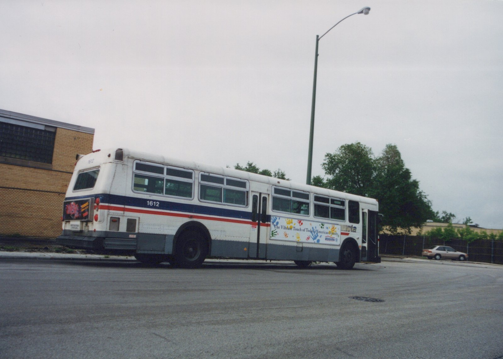 A white, blue, and red Chicago Transit Authority motor bus, number 1612, is parked on a street with a brick building and trees in the background under an overcast sky.