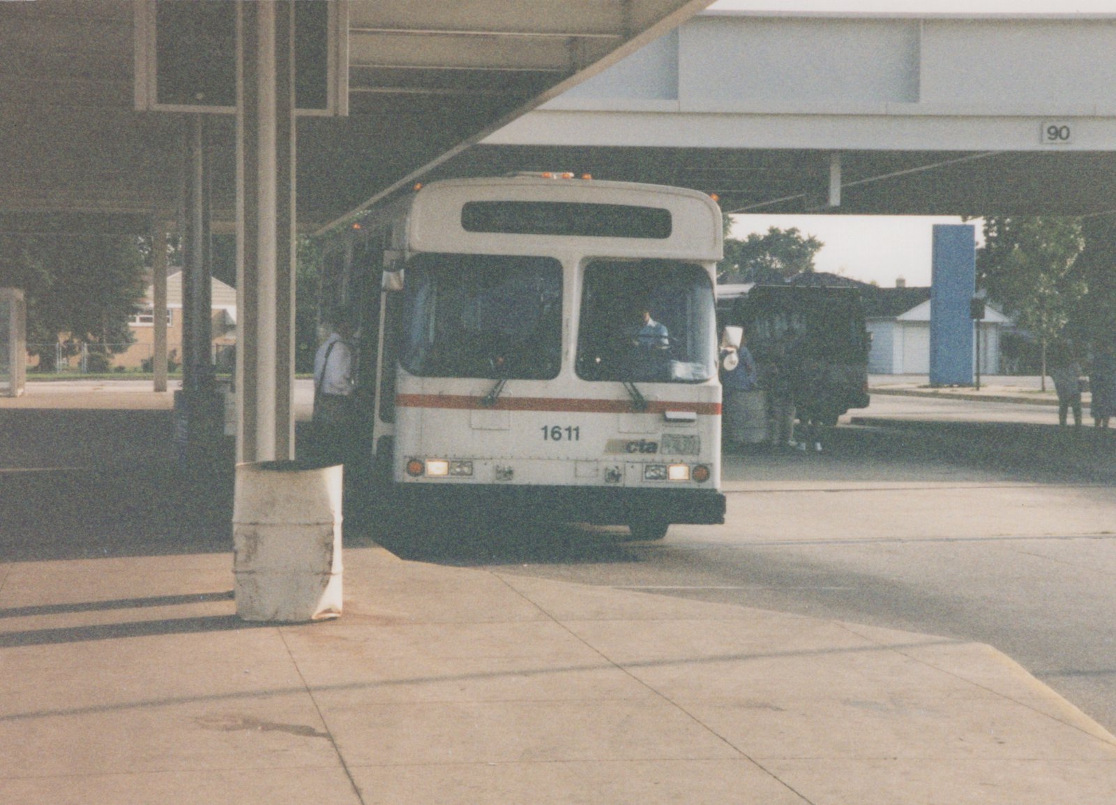 A white CTA bus with the number 1611 on the front, parked under a highway overpass, with a few people nearby.