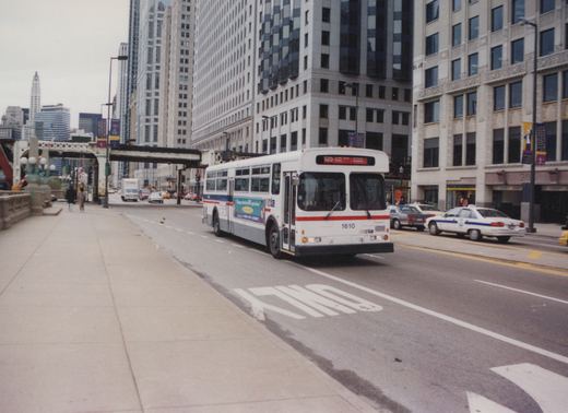 A white and blue CTA bus, number 1610, is shown on a city street with tall buildings and other vehicles in the background.