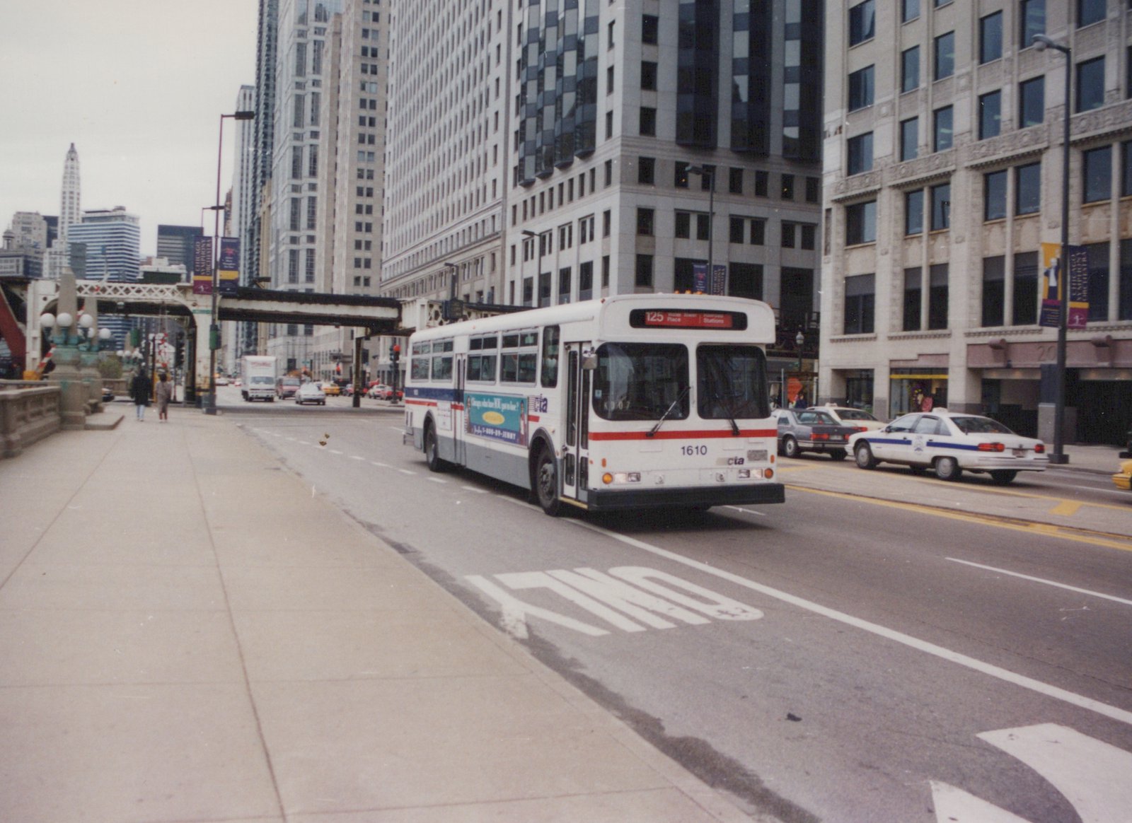 A white and blue CTA bus, number 1610, is shown on a city street with tall buildings and other vehicles in the background.