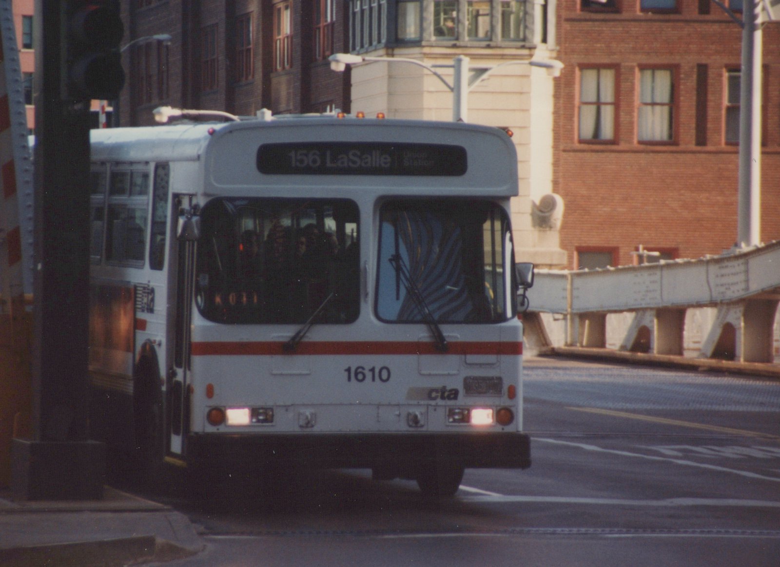 Front three-quarter view of a white and orange Chicago Transit Authority bus with number 1610 and destination 156 LaSalle Union Station, driving on a street with a bridge in the background.