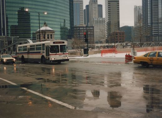 A white and blue CTA bus numbered 1608 drives on a wet city street with snow on the ground, surrounded by tall buildings and other vehicles, including a yellow taxi.