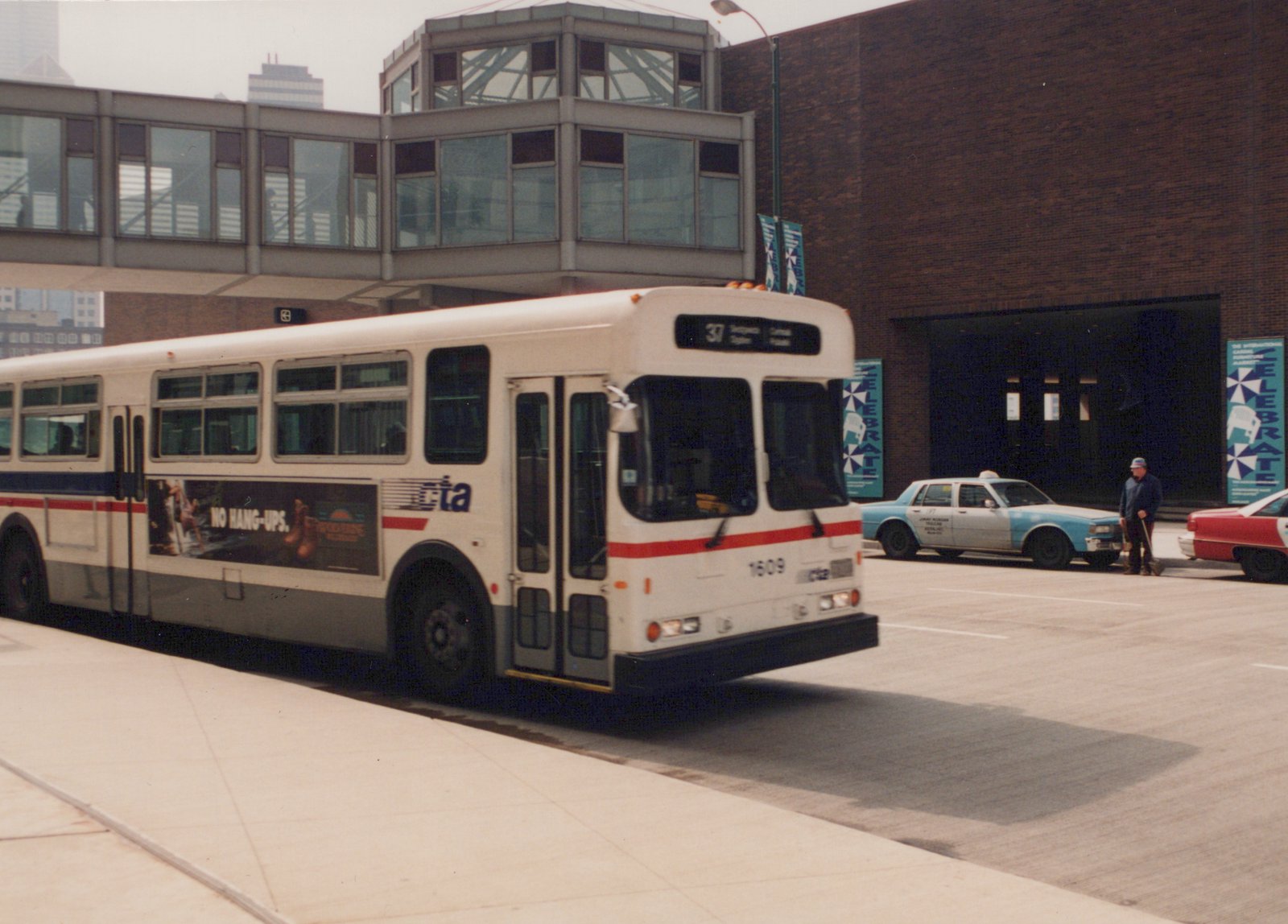 A white CTA bus with red and blue stripes, numbered 1609, is parked on a city street next to a brick building with a pedestrian bridge overhead and two taxis in the background.